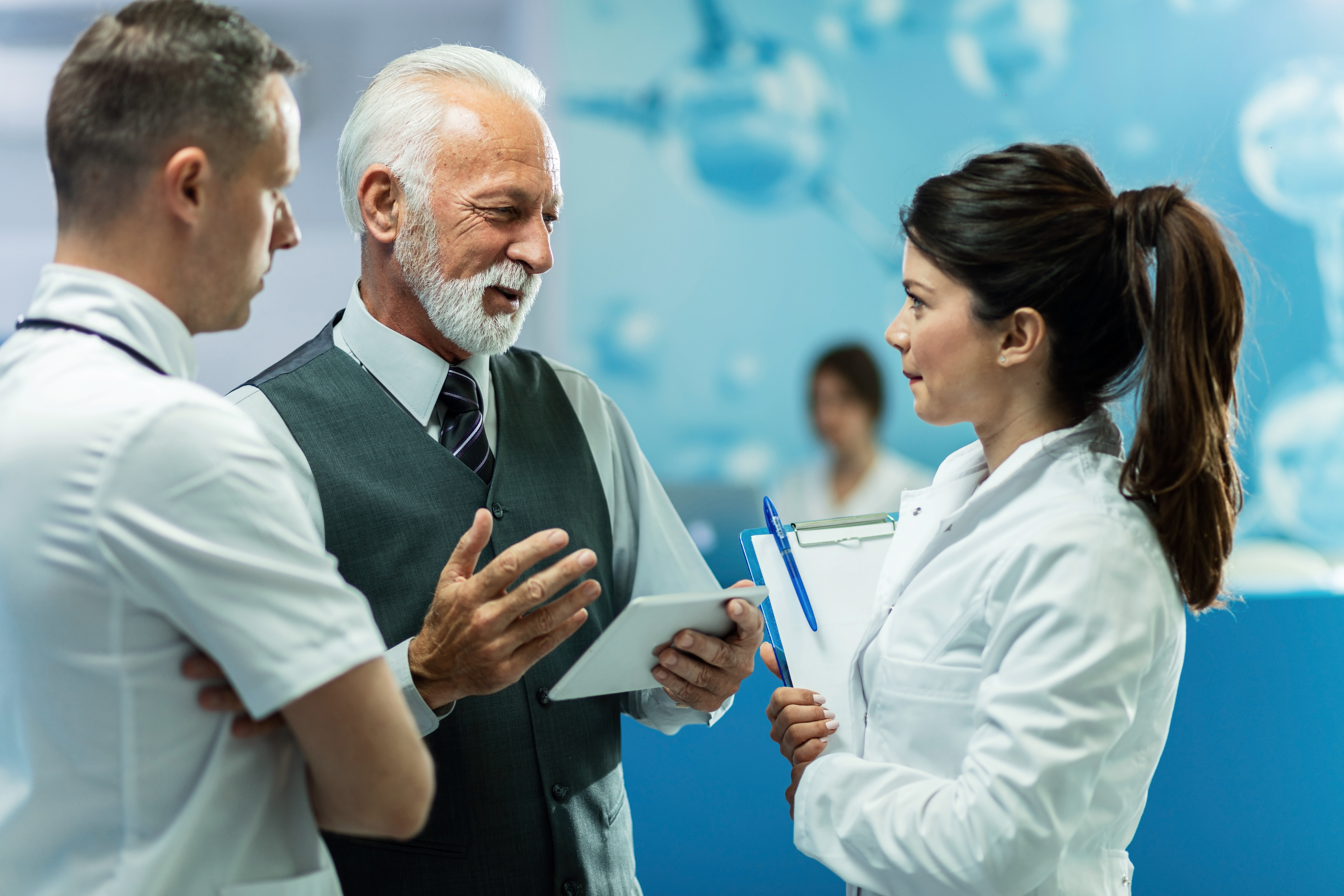 Happy mature businessman using digital tablet while talking to healthcare workers in a hallway at clinic.