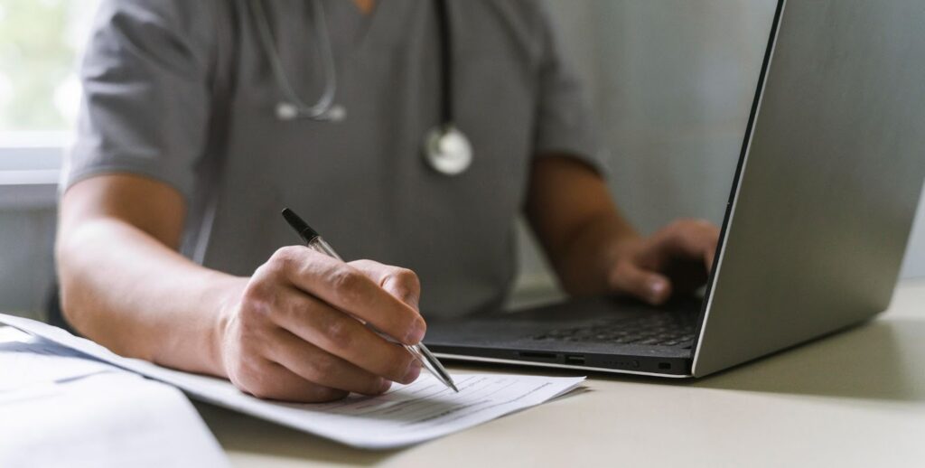 Side view of a doctor with a stethoscope working on a laptop and writing on paper